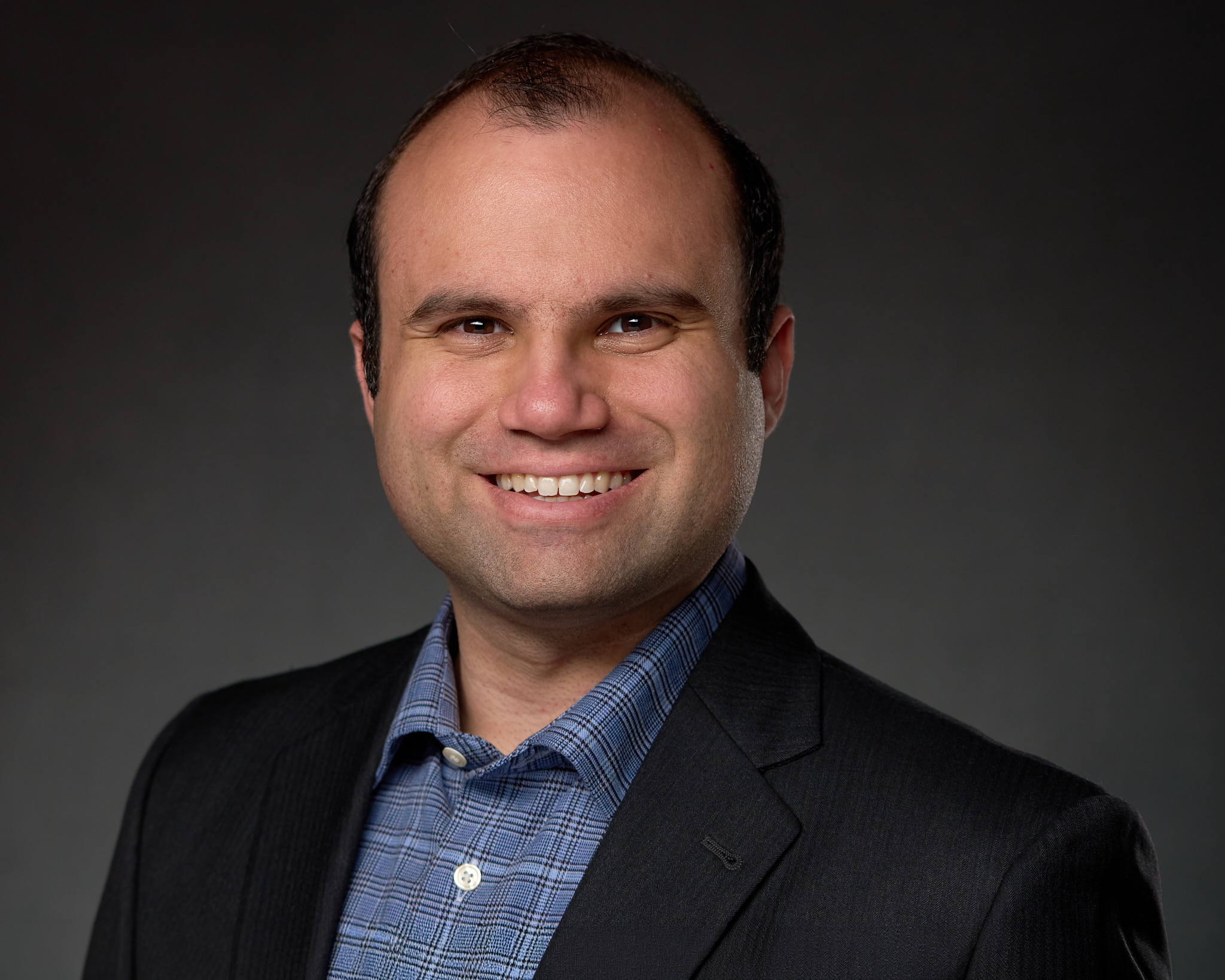 man with dark hair, strong catchlights in the eyes, big smile, wearing a sport coat over a blue tattersall dress shirt, in front of a gray background