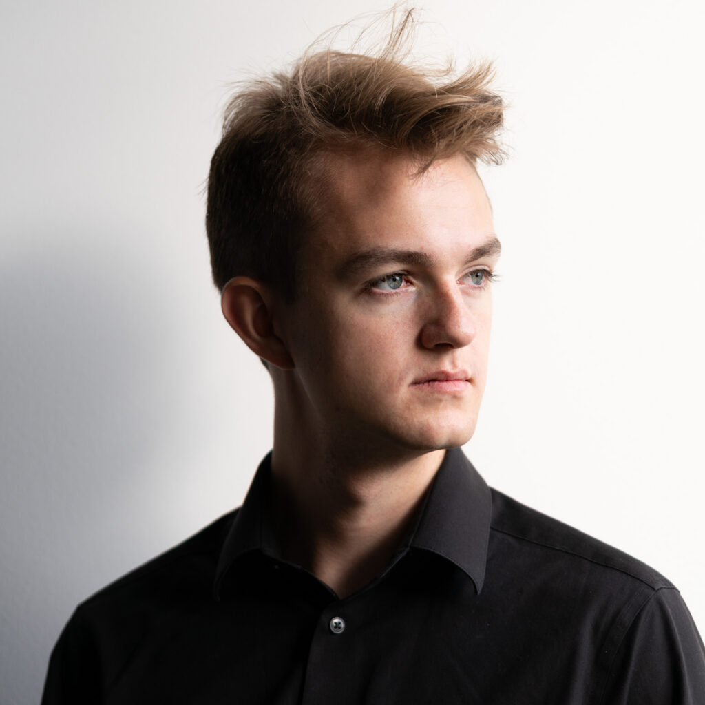 young man in front of white wall, looking to the right, neat brown hair with a flowing wave on top, black shirt, natural light