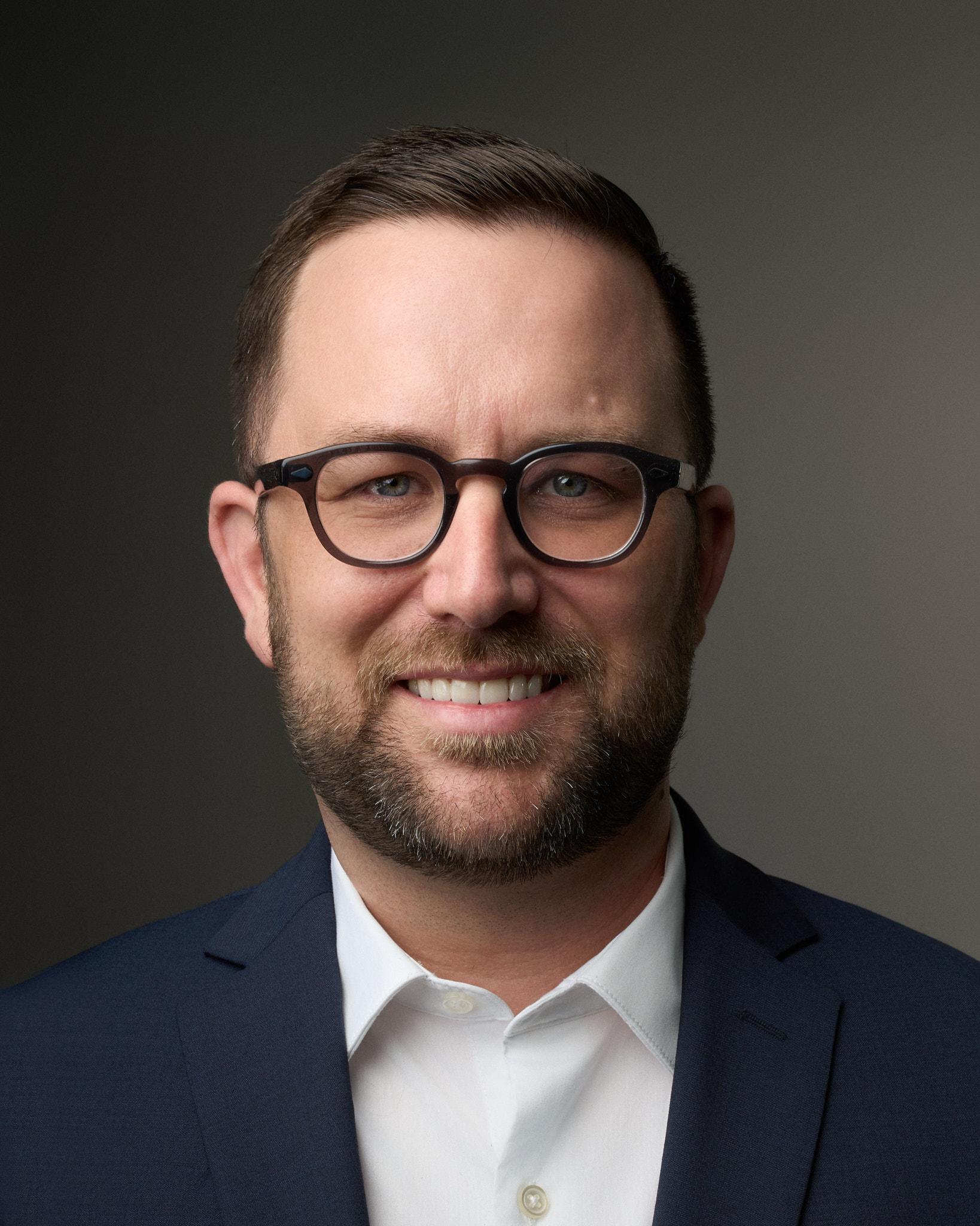 portrait of bearded man with neat brown hair, brown round-rimmed glasses, open smile, wearing blue sport coat and white dress shirt, background gray