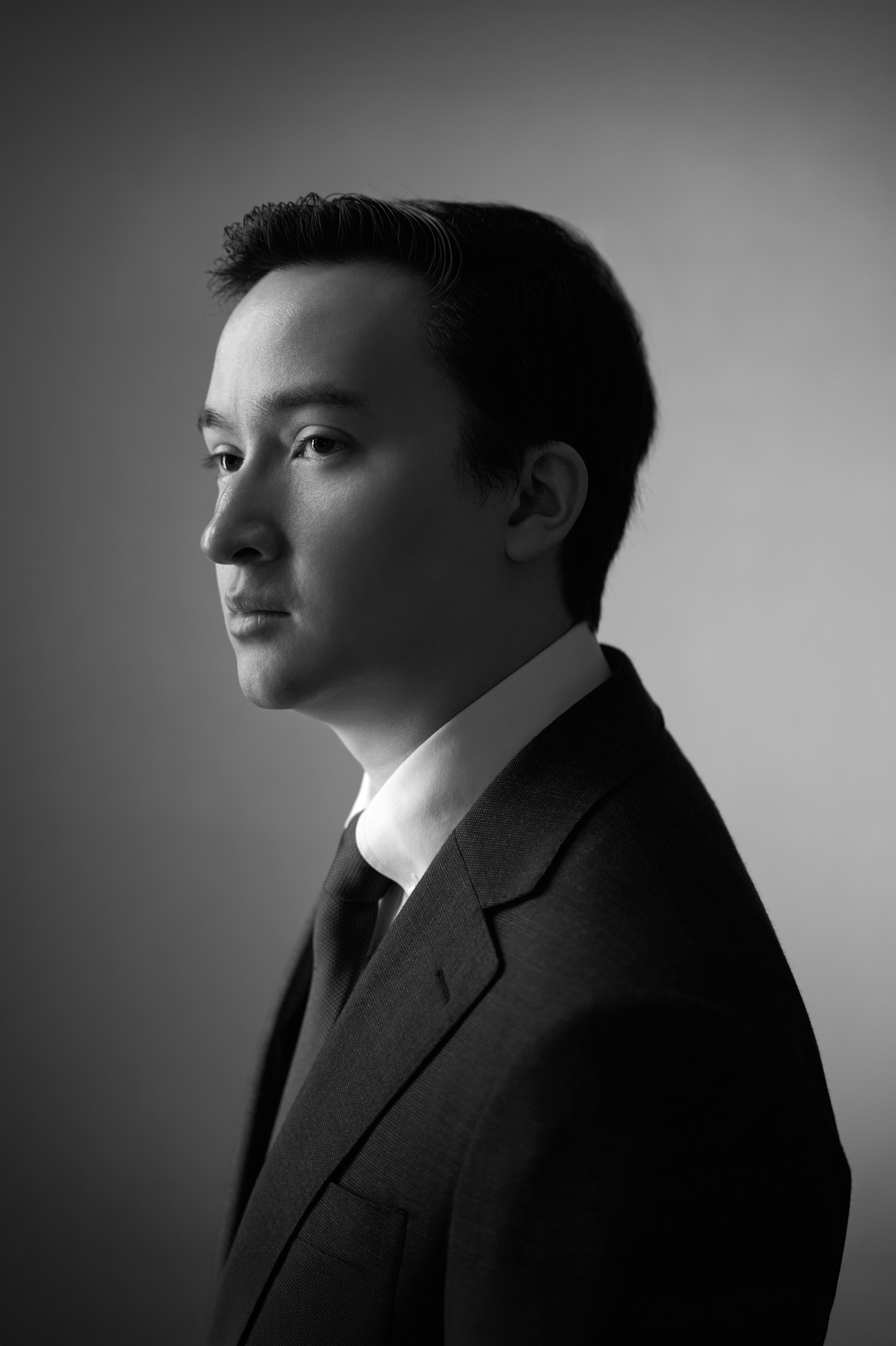 profile black and white image of young asian man, head turned slightly toward camera, serious expression, wearing a suit and tie, gradient background