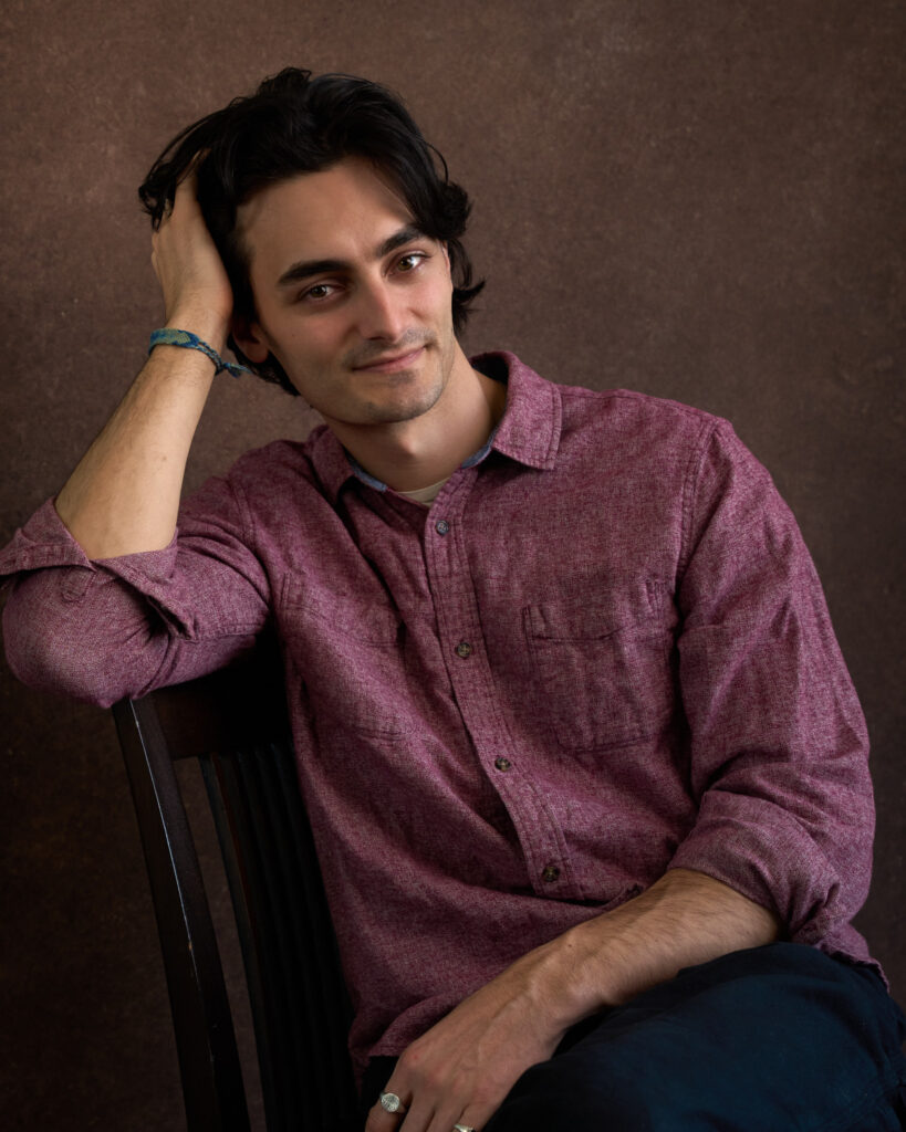 man seated with elbow on chair back, hand supporting head, red heather shirt, brown backdrop,